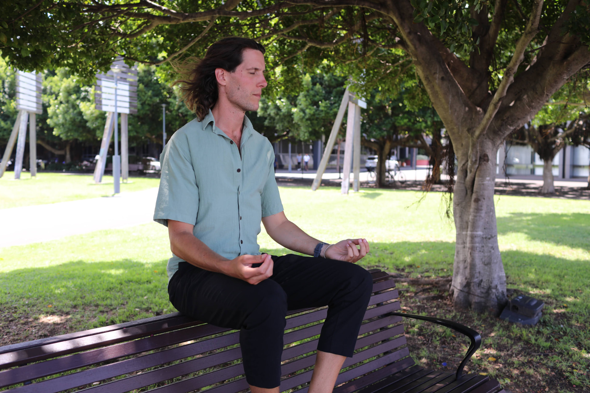 Sean sitting cross-legged on a park bench under a tree